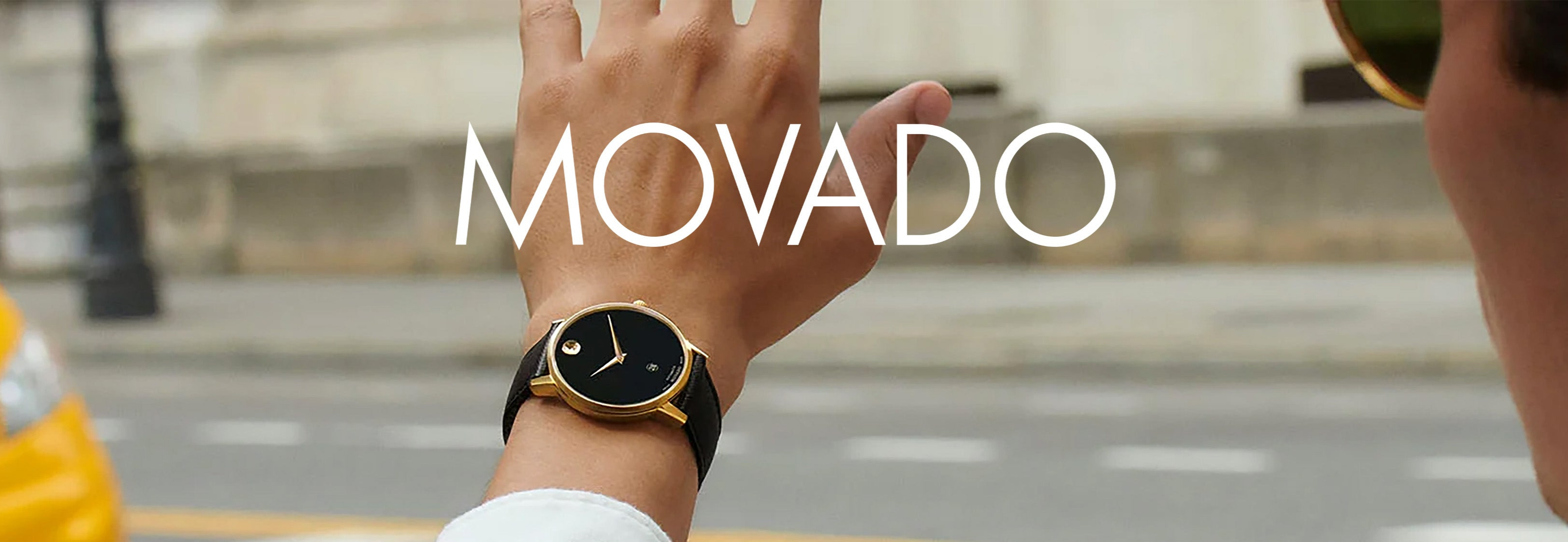 Luxury black and gold watch on wrist, street scene in background, MOVADO logo visible