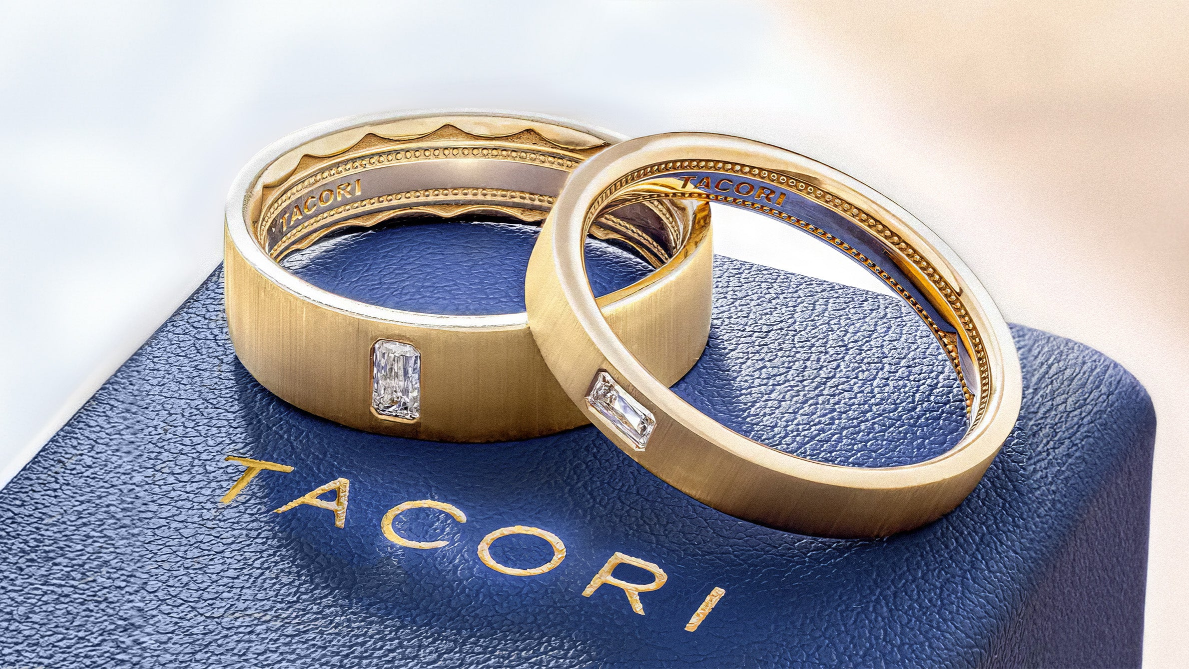 Gold wedding bands with inset diamonds on blue textured jewelry box, Tacori branding visible